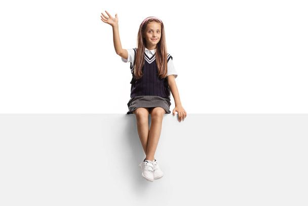 Schoolgirl with long hair sitting on a blank panel and waving isolated on white background - Фото, зображення