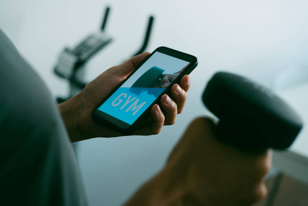 крупним планом молодий кавказький чоловік піднімає гантелі у вітальні свого будинку, дивлячись на свій смартфон, який читає спортзал на екрані, і стаціонарний велосипед на задньому плані
 - Фото, зображення