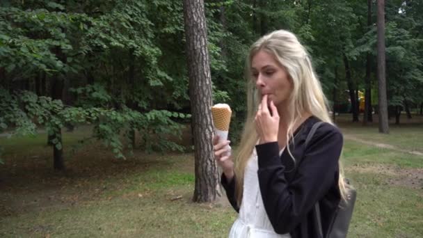 Ein junges schönes Mädchen in weißem Kleid und schwarzer Strickjacke im Park und isst eine Waffel mit Eis und lächelt sehr emotional - Filmmaterial, Video