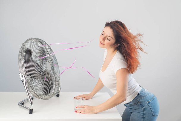A red-haired Caucasian woman chills by the electric fan and drinks a cold drink. Climate control in the apartment - Foto, afbeelding