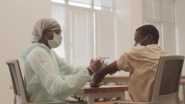 Średnie ujęcie pracownicy służby zdrowia w odzieży ochronnej wykonującej wstrzyknięcia młodemu Afroamerykaninowi szczepionemu przeciwko Covid-19 - Materiał filmowy, wideo