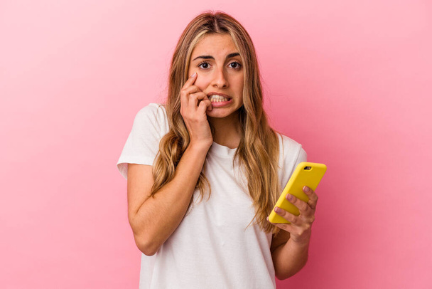 Молодая блондинка белая женщина, держащая желтый мобильный телефон изолирован грызет ногти, нервный и очень тревожный. - Фото, изображение
