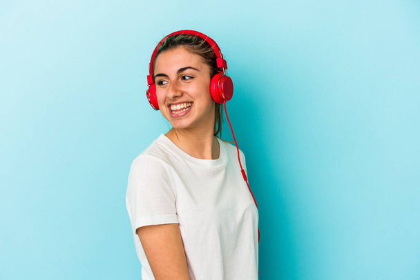 Young blonde woman listening to music on headphones isolated on blue background looks aside smiling, cheerful and pleasant. - Fotó, kép