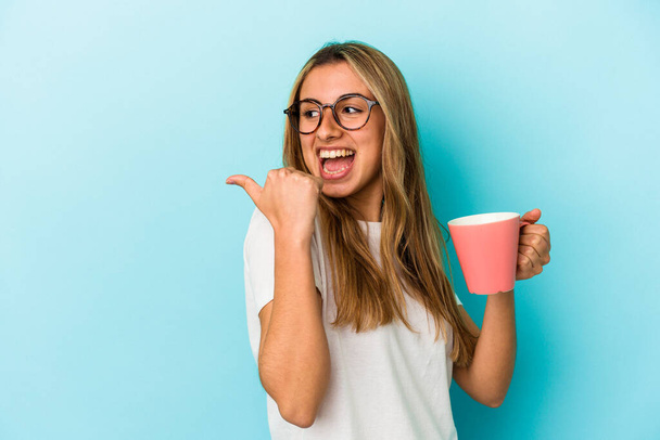 Young caucasian blonde woman holding a mug isolated on blue background points with thumb finger away, laughing and carefree. - Foto, immagini