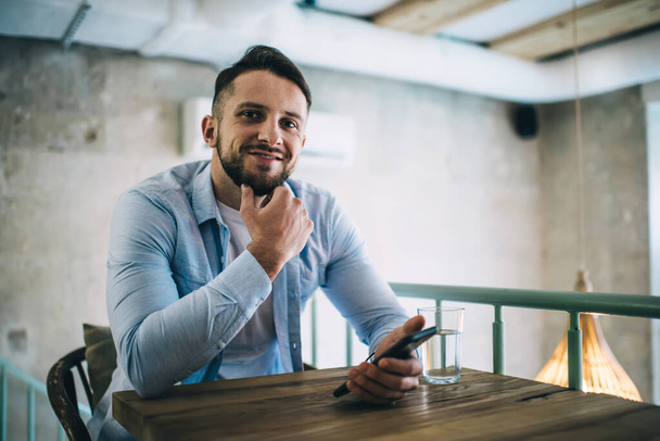 Portrait of smiling caucasian male blogger enjoying spending free time at cafeteria using wireless connection on smartphone for chatting, handsome millennial hipster guy using modern mobile phone - 写真・画像