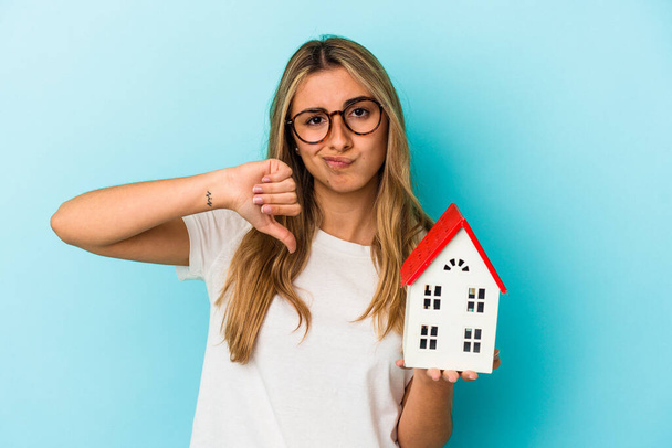 Young caucasian woman holding a house model isolated on blue background showing a dislike gesture, thumbs down. Disagreement concept. - Фото, изображение