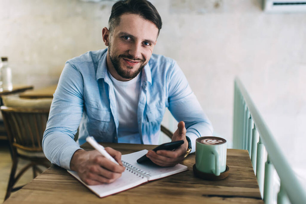 Smiling caucasian male writer using smartphone for making research in social networks sitting in cafe look at camera, portrait of prosperous male blogger writing article in notebook in coffee shop - Photo, image