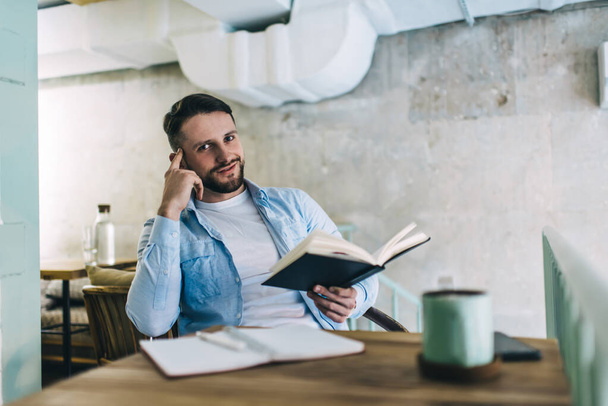 Portrait of handsome smiling male student enjoying learning sitting at cafe interior with books for research,smiling hipster guy spending time on education spending time in coffee shop with literature - Фото, изображение