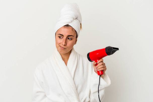 Mulher branca jovem segurando um secador de cabelo isolado no branco confuso, se sente duvidoso e inseguro. - Foto, Imagem