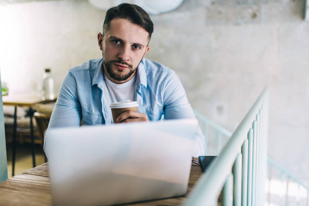 Portrait of serious caucasian millennial freelancer working remotely holding disposable cup with beverage, confident businessman sitting at laptop computer for checking mail and browsing in cafe - Photo, image