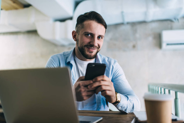 Portrait of prosperous male freelancer satisfied with earning online checking making account and transaction on smartphone, happy hipster guy student learning using modern devices in coffee shop - Photo, Image