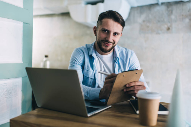 Half length portrait of handsome male entrepreneur checking data and income from coffee shop holding planner, positive hipster guy freelancer looking at camera making research in cafe with laptop - Фото, изображение