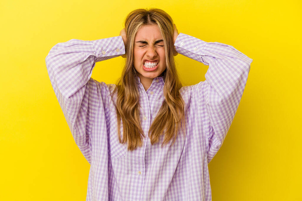 Young blonde caucasian woman isolated on yellow background covering ears with hands trying not to hear too loud sound. - Photo, image