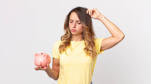 pretty thin woman feeling puzzled and confused, scratching head and holding a piggybank - Foto, Imagem
