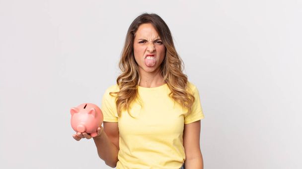 pretty thin woman feeling disgusted and irritated and tongue out and holding a piggybank - Foto, Imagem