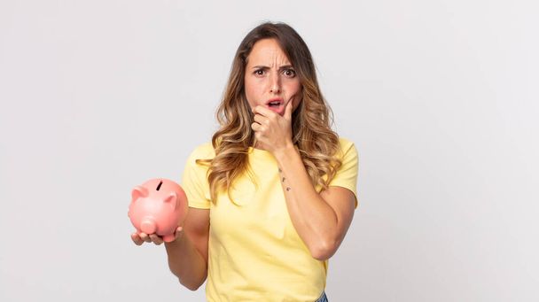 pretty thin woman with mouth and eyes wide open and hand on chin and holding a piggybank - 写真・画像