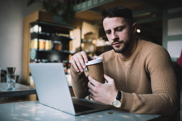Handsome bearded hipster guy in casual wear looking at disposable coffee cup while working via laptop computer, pensive male student learning in cafe interior online enjoy beverage in morning - Fotoğraf, Görsel