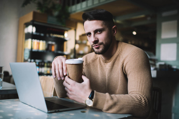 Serious handsome male entrepreneur holding coffee cup looking at camera during remote job process, caucasian 20s businessman enjoying coffee while having freelance project on laptop computer - Φωτογραφία, εικόνα