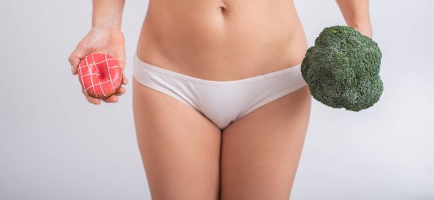Uma mulher sem rosto de calcinha branca segura um donut e brócolis em um fundo branco. Hábitos alimentares. - Foto, Imagem