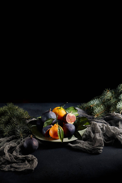 Dark Mood Kerst Samenstelling met Fruit Plate en Christmas Tree Branch. Vintage tafel met paarse figuren en mandarijn op donkergroene plaat - Foto, afbeelding