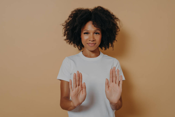 Młoda uśmiechnięta mieszanka rasy kobieta z kręconą fryzurą podnoszenie dłoni w stop lub zakaz gestu - Zdjęcie, obraz