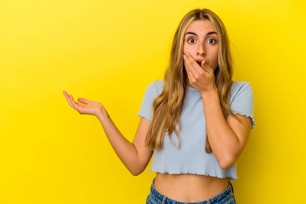 Young blonde caucasian woman isolated on yellow background holds copy space on a palm, keep hand over cheek. Amazed and delighted. - Foto, immagini