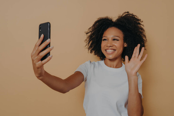 陽気なアフリカ人女性ジェスチャーhiしながらビデオ通話をしたり、スマートフォンで自画撮り - 写真・画像