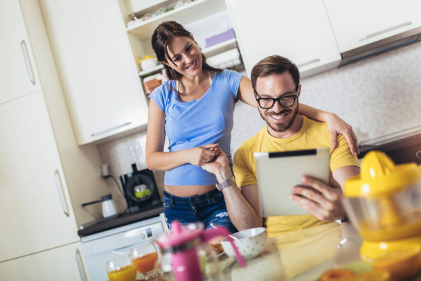 Terhes nő férjjel a konyhában szórakozik. digitális tabletta használata. - Fotó, kép