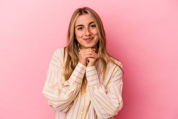Young blonde caucasian woman isolated on pink background keeps hands under chin, is looking happily aside. - Photo, Image