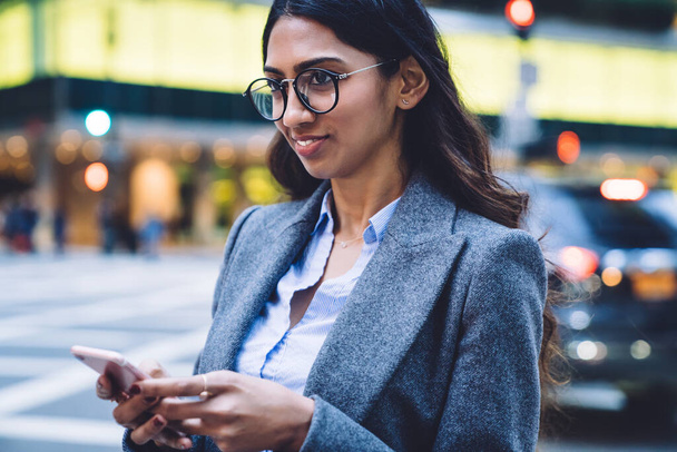 Young pretty ethnic business woman in gray jacket holding mobile phone and thinking while standing outdoors in New York City - Foto, afbeelding