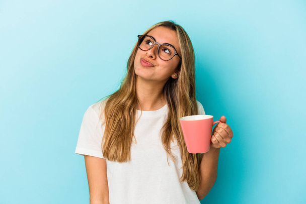 Young caucasian blonde woman holding a mug isolated on blue background dreaming of achieving goals and purposes - Foto, afbeelding