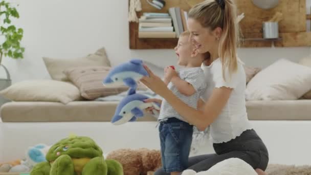 Gelukkig en lachende moeder met baby spelen in de woonkamer thuis met pluche knuffels dieren speelgoed, zitten op de vloer met kussens, Kaukasische blonde met blauwe ogen, gezond en liefde leven groei concept - Video
