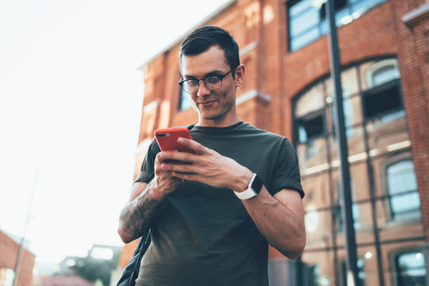 From below of content male blogger in stylish casual clothing and trendy eyeglasses with smartwatch on wrist chatting in mobile phone at city street  - 写真・画像