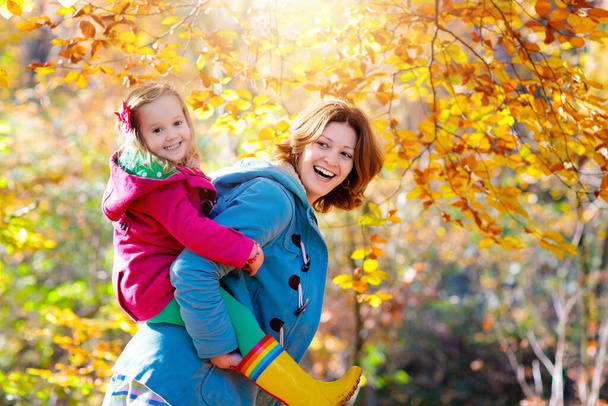 Matka i dziecko bawią się w jesiennym parku. Mama i dziecko spacerują po lesie w słoneczny jesienny dzień. Dzieci bawiące się na zewnątrz z żółtym liściem klonu. Maluch i rodzic zbierają złote liście. Mama przytula dziecko. - Zdjęcie, obraz