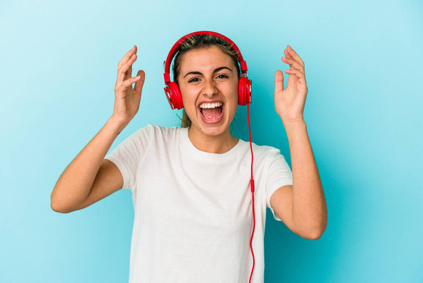 Junge blonde Frau hört Musik über Kopfhörer isoliert auf blauem Hintergrund und erhält eine angenehme Überraschung, ist aufgeregt und hebt die Hände. - Foto, Bild