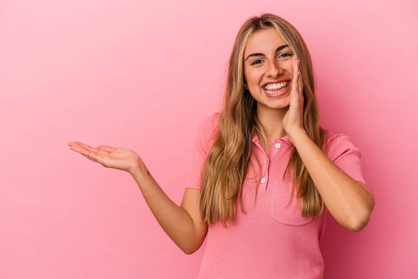La donna caucasica bionda giovane isolata su sfondo rosa tiene lo spazio di copia su una palma, tenga la mano sopra guancia. Stupito e felice. - Foto, immagini