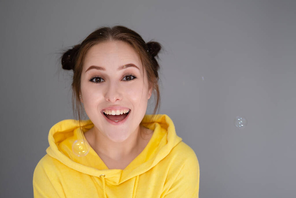 cheerful positive brunette woman in yellow hoodie in white bedroom on bed with white linens. happy people. millennial generation. fashionable teenager. - Photo, Image