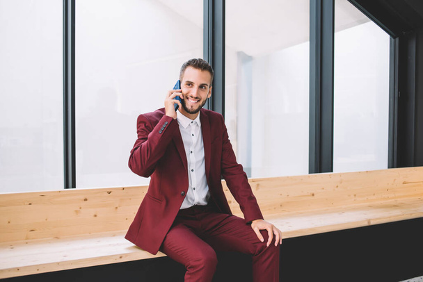 Joyful ethnic guy in formal suit chatting with friend using cellphone resting from freelance work in office room looking away - Foto, immagini