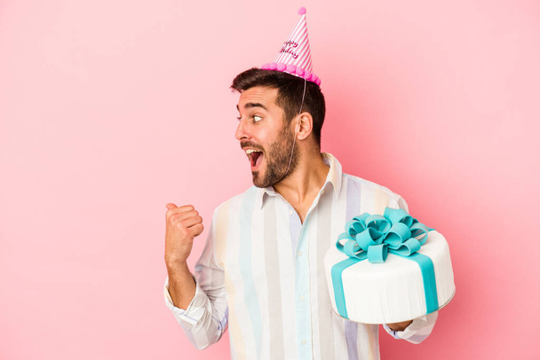 Young caucasian man celebrating his birthday isolated on pink background points with thumb finger away, laughing and carefree. - 写真・画像