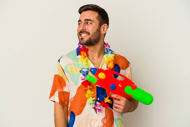 Young caucasian woman on a hawaiian party holding a water gun isolated on white background looks aside smiling, cheerful and pleasant. - Foto, Imagen