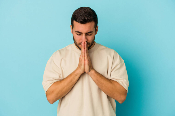 Young caucasian man isolated on blue background praying, showing devotion, religious person looking for divine inspiration. - Fotografie, Obrázek