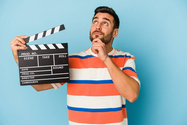 Young caucasian man holding a clapperboard isolated on blue background looking sideways with doubtful and skeptical expression. - Photo, Image