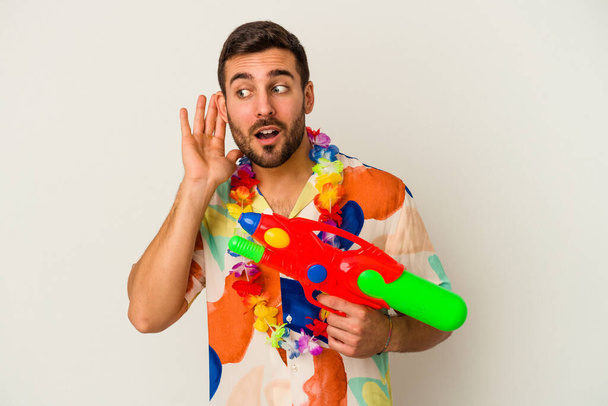 Young caucasian woman on a hawaiian party holding a water gun isolated on white background trying to listening a gossip. - Foto, afbeelding