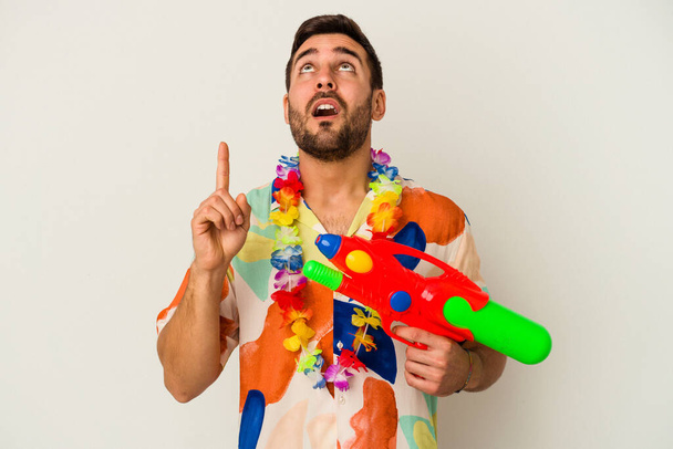 Young caucasian woman on a hawaiian party holding a water gun isolated on white background pointing upside with opened mouth. - Foto, imagen