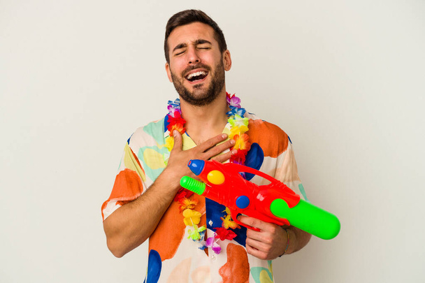 Молодая кавказка на гавайской вечеринке, держащая водяной пистолет на белом фоне, громко смеется, держа руку на груди.. - Фото, изображение