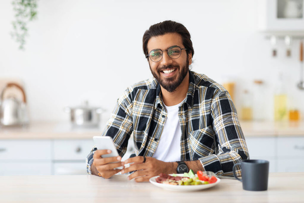 Χαρούμενος μεσανατολίτης άνθρωπος απολαμβάνοντας πρωινό, κρατώντας smartphone - Φωτογραφία, εικόνα