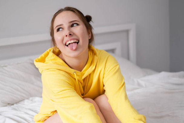 cheerful positive brunette woman in yellow hoodie in white bedroom on bed with white linens. happy people. millennial generation. fashionable teenager. - Photo, Image