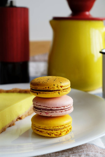 Žluté a růžové makrónky hromádka na bílém talíři v kávové zátiší. Ruční vaření, žlutý keramický džbán, červená mlýnek na kávu - Fotografie, Obrázek