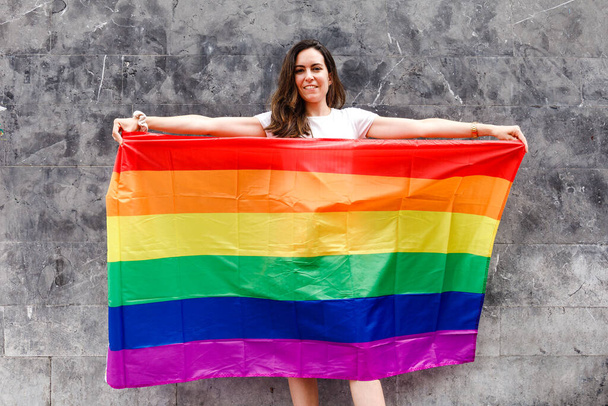 Mujer joven sosteniendo bandera de arco iris sobre fondo gris - Foto, imagen