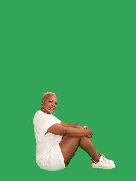 Vertical portrait of a smiling beautiful Afro American woman wearing sportswear seating isolated against a green background - Foto, Bild